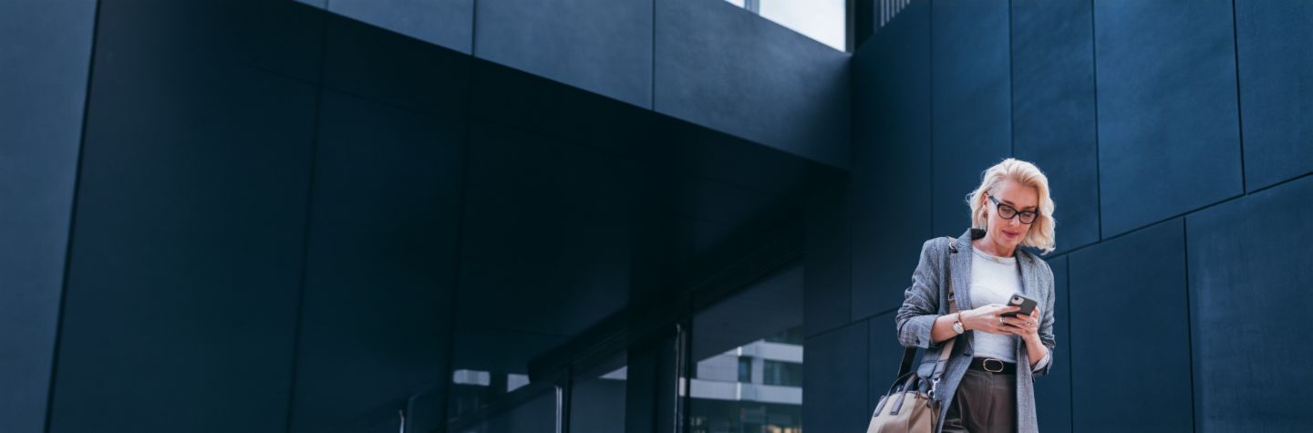 Een blonde zakenvrouw in een grijze blazer staat voor een modern gebouw en kijkt glimlachend op haar smartphone.