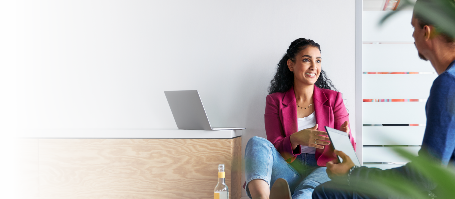 Twee jonge vrouwen, de linker in een roze blazer met zwarte broek en de rechter in een wit shirt met beige broek, lopen naast elkaar voor een witte muur in een licht gebouw. Ze praten met elkaar. Op de voorgrond, aan de rechterkant, zien we een vaag deel van een groene plant.