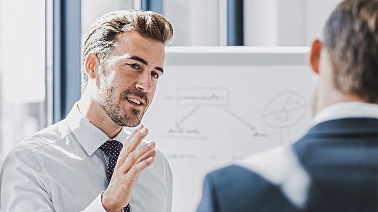 On voit un homme en tenue professionnelle expliquer quelque chose avec une expression amicale et engagée. Face à lui se tient une autre personne qui l’écoute attentivement. On aperçoit en arrière-plan un tableau blanc avec un croquis.