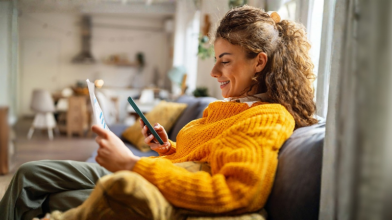 Une jeune femme souriante aux cheveux bouclés, vêtue d'un pull jaune confortable, est assise sur un canapé dans son salon et regarde son smartphone tout en tenant un document papier.