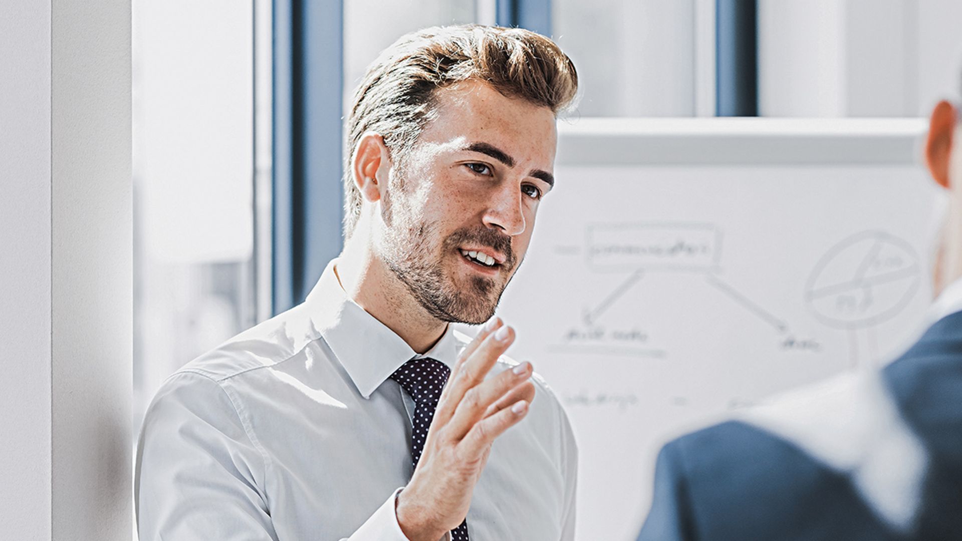 Je ziet een man in zakelijke kleding die met een vriendelijke, betrokken uitdrukking iets uitlegt. Voor hem staat een andere persoon die duidelijk naar hem luistert. Op de achtergrond is een whiteboard met een schets te zien.