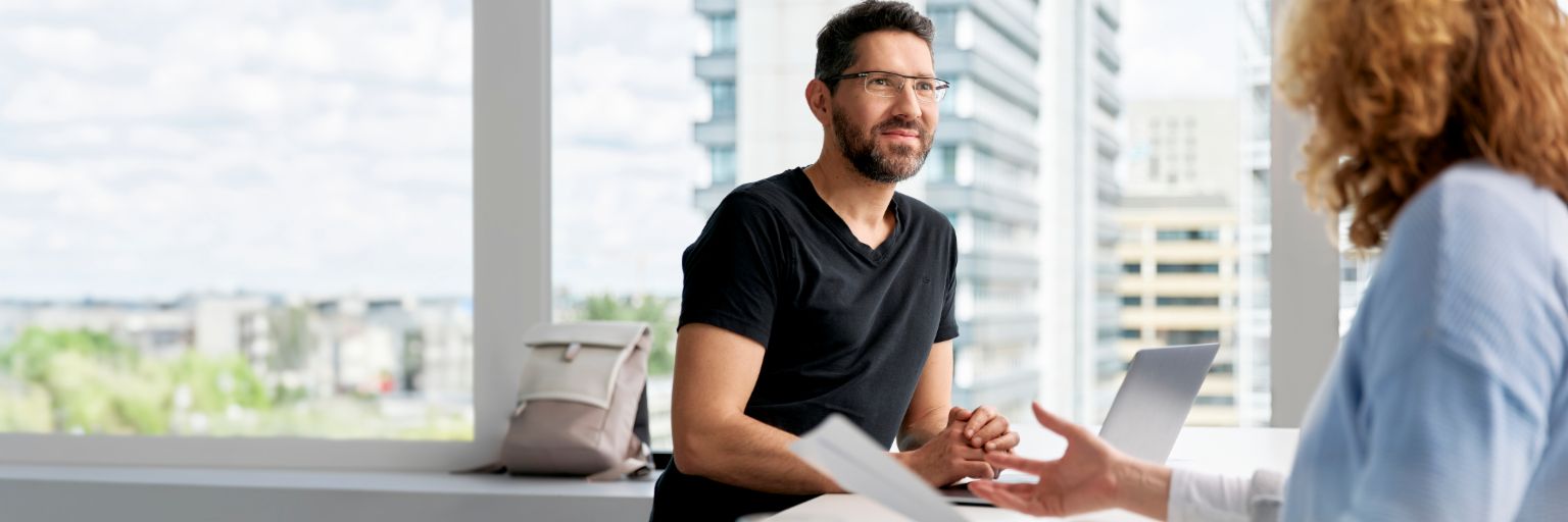 Twee mensen praten met elkaar in een moderne, lichte kantoorruimte. <br/>In het midden van de foto glimlacht een man met donker haar, een bril en een baard. Hij draagt een zwart T-shirt. Op de tafel voor hem liggen een paar documenten en een laptop. De tweede persoon, van wie de armen en het hoofd in beeld zijn, zit tegenover de man. Ze draagt een lichtblauwe bloes, heeft kort, krullend, roodblond haar en heeft een paar vellen papier vast. Op de achtergrond zien we grote ramen, met daarachter moderne gebouwen in een stad.