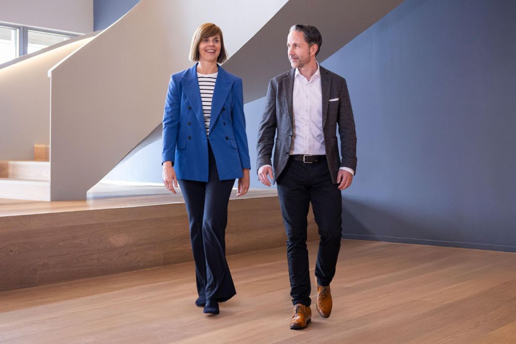 Le PDG en blazer gris et le CFO du Groupe EOS en blazer bleu traversent un bureau moderne. L'espace dispose de grandes fenêtres, d'un mur courbé et de lampes suspendues au design élégant.