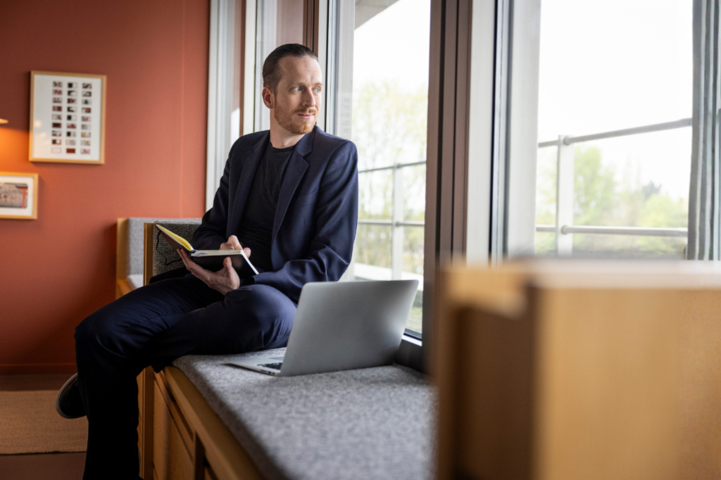 Sebastian Pollmer zit op een vensterbank met een open laptop en een notitieboek in zijn hand, terwijl hij uit het raam staart.
