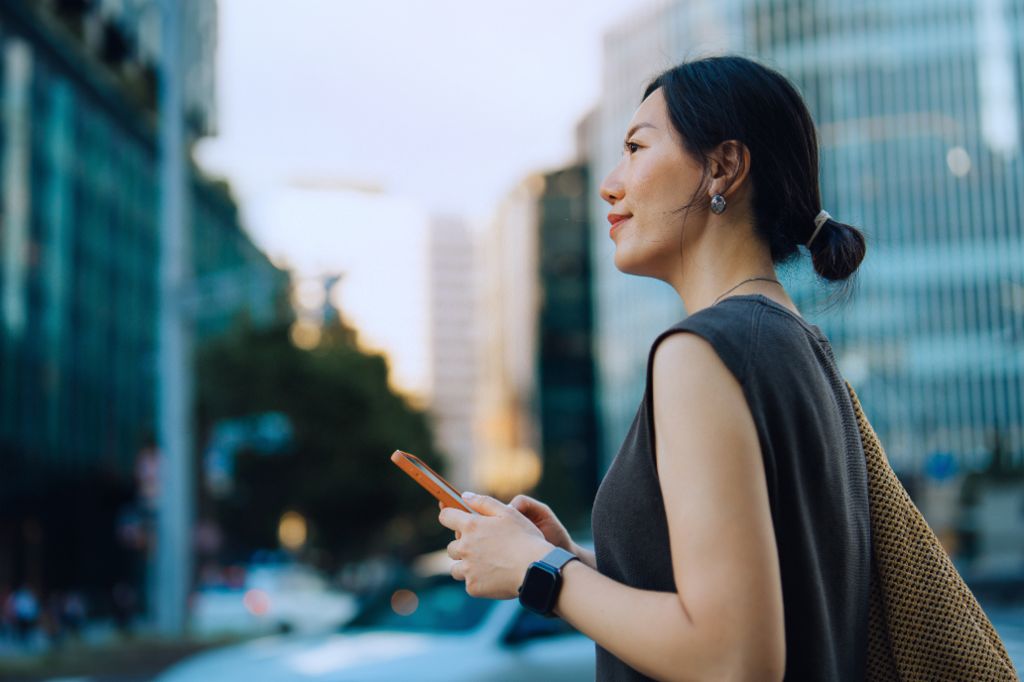 Een jonge vrouw staat buiten in een stedelijke omgeving, houdt haar smartphone vast en kijkt glimlachend vooruit. Op de achtergrond zijn moderne gebouwen en bomen te zien.