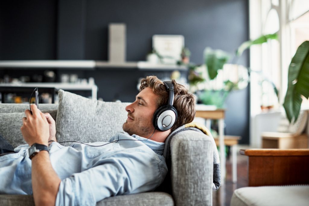 Een jonge man ligt op een bank in zijn woonkamer, draagt een koptelefoon en kijkt lachend naar zijn smartphone terwijl hij geniet van muziek of een podcast.