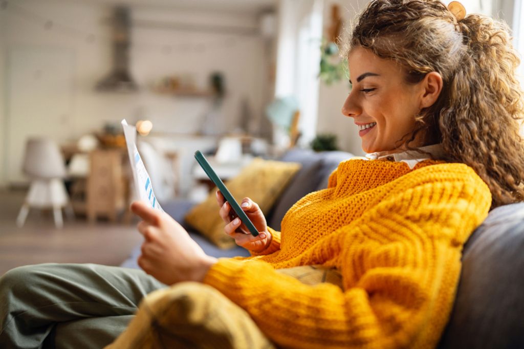 Een lachende jonge vrouw met krullend haar, gekleed in een gezellige gele trui, zit op de bank in haar woonkamer en kijkt op haar smartphone terwijl ze een papieren document vasthoudt.