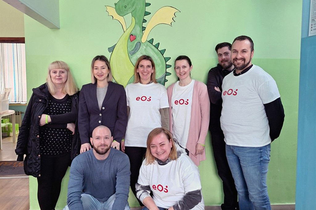 Acht mensen staan voor een muur waarop een groene draak is geschilderd. Ze staan in twee rijen: twee zitten gehurkt vooraan en zes staan achteraan. Vier mensen dragen een witte T-shirt met het rode EOS-logo. De groep lacht naar de camera. Op de vloer, vooraan rechts, staan een paar flessen en drankkartons.