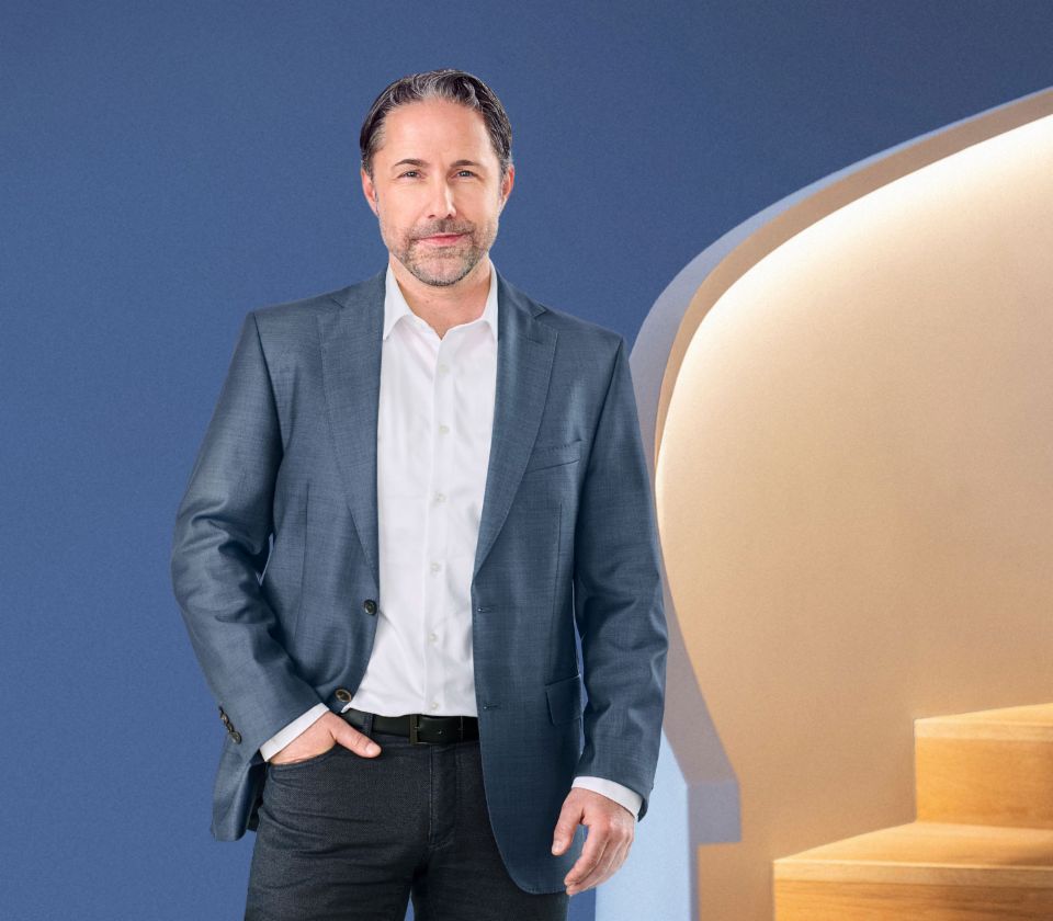 Marwin Ramcke, Chief Executive Officer van de EOS Group, staat voor een blauwe achtergrond, gekleed in een donker jasje. Hij kijkt recht in de camera.