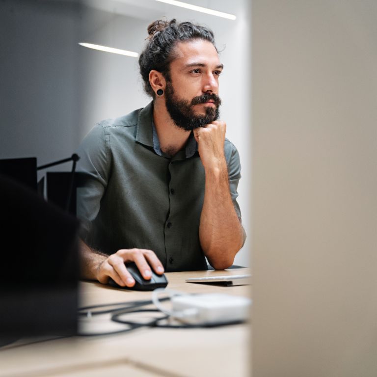 Een man met een baard en lang haar in een knot zit achter een computer, volledig verdiept in zijn werk.