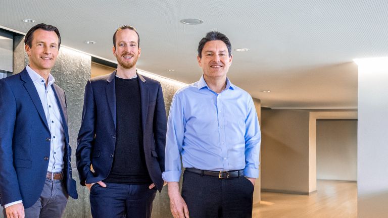 Drie mannelijke leden van de Raad van Bestuur van de EOS Groep staan in zakelijke kleding voor liften in een moderne omgeving.