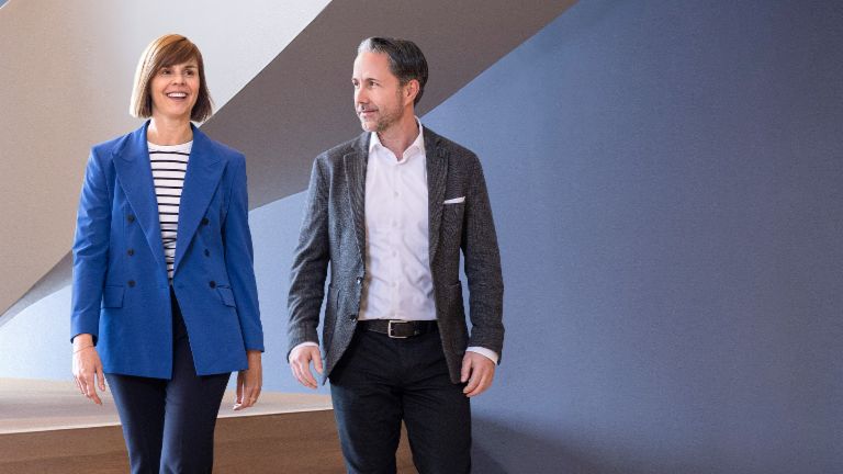 De CEO in een grijze blazer en de CFO van de EOS Groep in een blauwe blazer lopen door een modern kantoor. De ruimte heeft grote ramen, een gebogen muur en stijlvolle hanglampen.