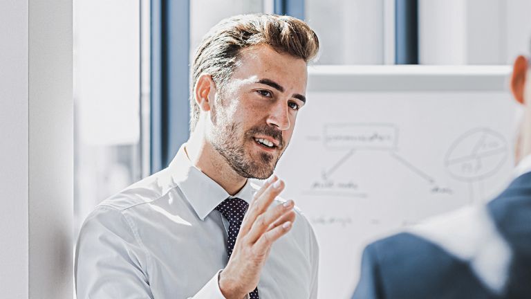 Je ziet een man in zakelijke kleding die met een vriendelijke, betrokken uitdrukking iets uitlegt. Voor hem staat een andere persoon die duidelijk naar hem luistert. Op de achtergrond is een whiteboard met een schets te zien.