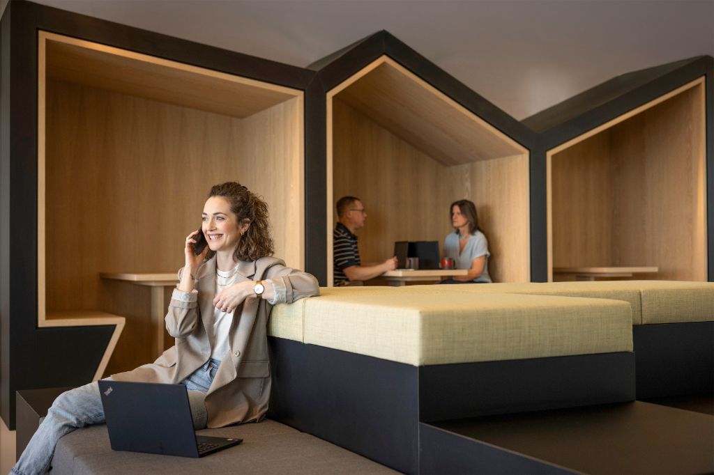 Een glimlachende persoon zit in een moderne kantoorlounge, telefoneert en werkt op een laptop. Op de achtergrond zijn twee andere mensen aan het werk.