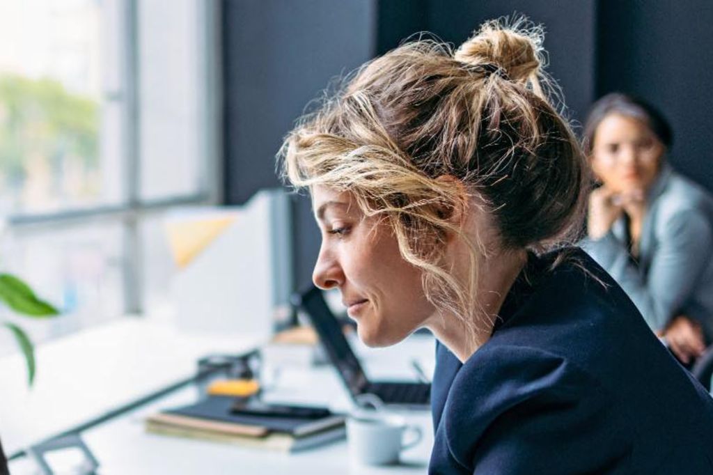 De afbeelding toont een vrouw die aan een bureau zit in een modern kantoor, gekleed in een donker blazer terwijl ze zich concentreert op haar laptop, met een plant en enkele kantoorbenodigdheden zichtbaar op de tafel, en een andere persoon die op de achtergrond zit.