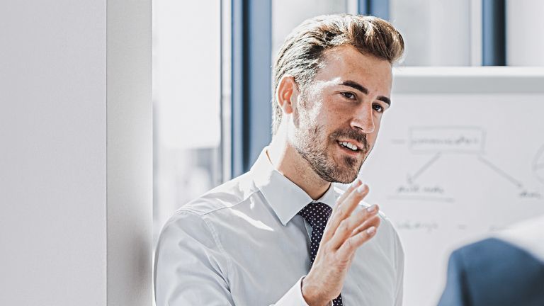 On voit un homme en tenue professionnelle expliquer quelque chose avec une expression amicale et engagée. Face à lui se tient une autre personne qui l’écoute attentivement. On aperçoit en arrière-plan un tableau blanc avec un croquis.