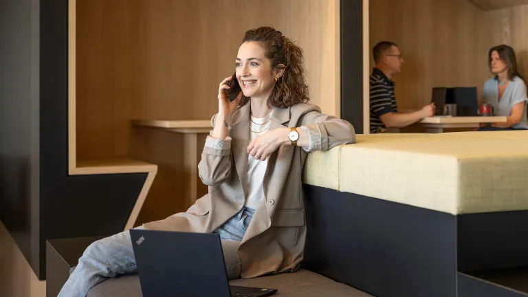 Une personne souriante est assise dans un salon de bureau moderne, parle au téléphone et travaille sur un ordinateur portable. En arrière-plan, deux autres personnes travaillent.