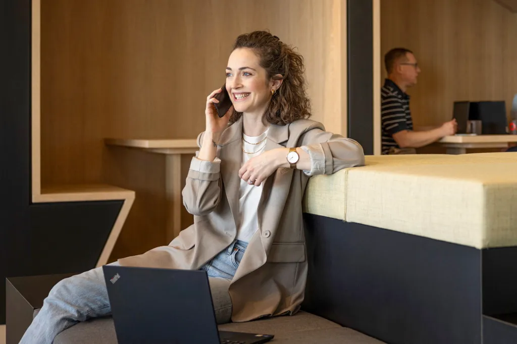 Une personne souriante est assise dans un salon de bureau moderne, parle au téléphone et travaille sur un ordinateur portable. En arrière-plan, deux autres personnes travaillent.