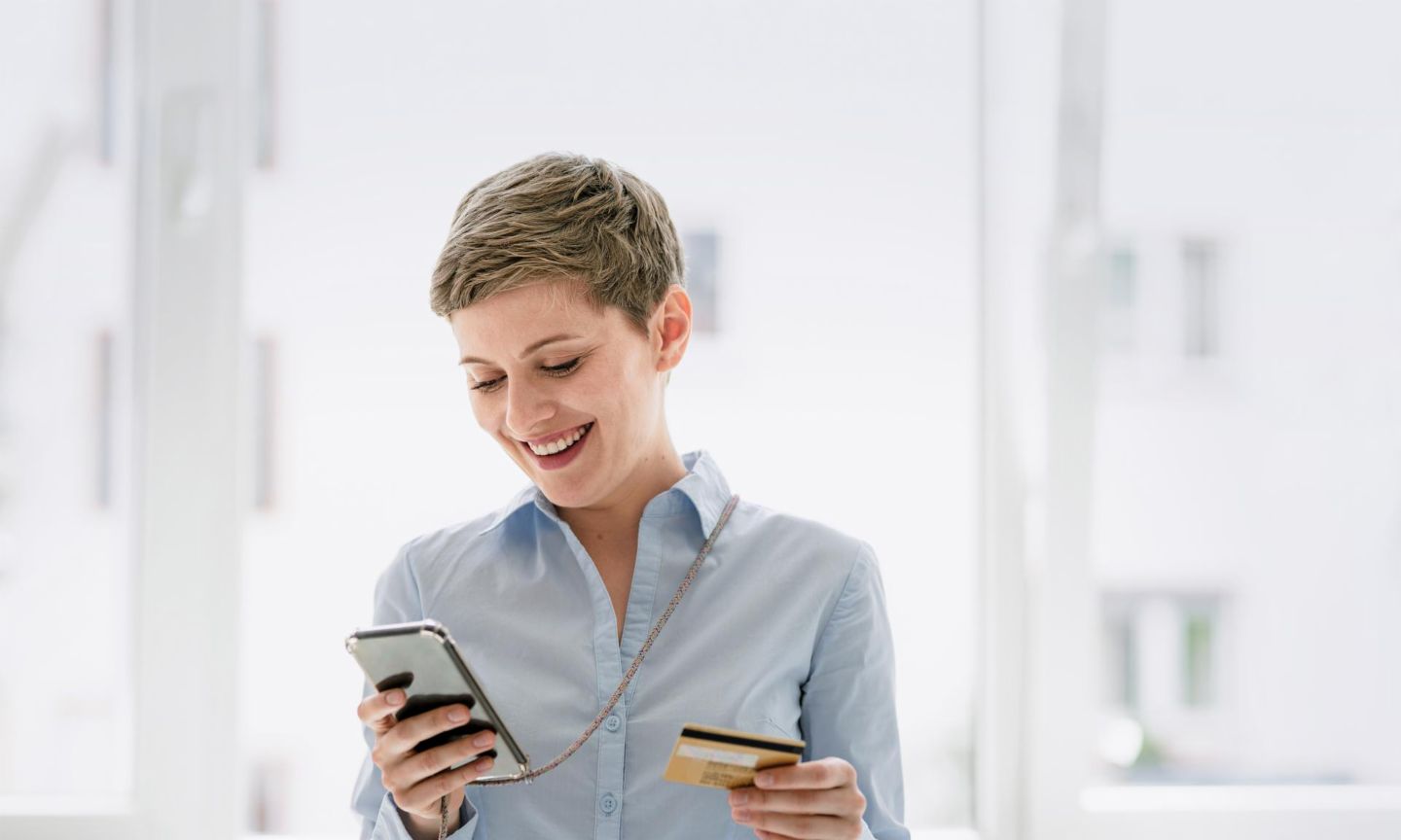Woman with short hair looks at her mobile phone and has her credit card in the other hand. 