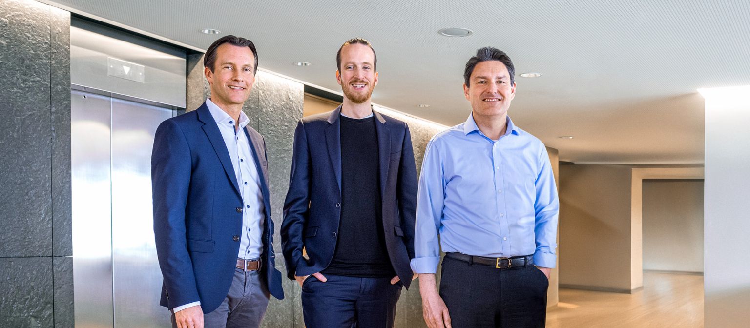 Drie mannelijke leden van de Raad van Bestuur van de EOS Groep staan in zakelijke kleding voor liften in een moderne omgeving.