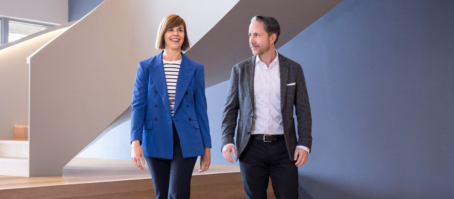 De CEO in een grijze blazer en de CFO van de EOS Groep in een blauwe blazer lopen door een modern kantoor. De ruimte heeft grote ramen, een gebogen muur en stijlvolle hanglampen.