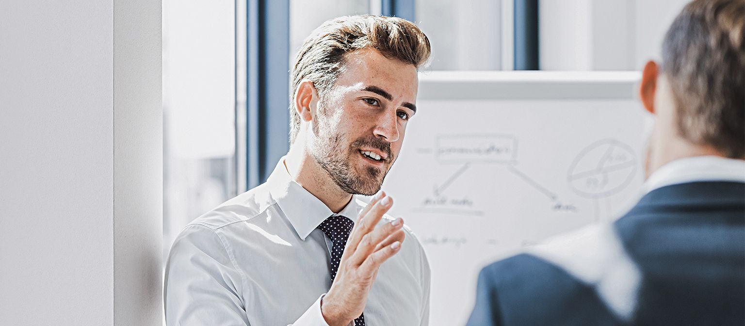 Je ziet een man in zakelijke kleding die met een vriendelijke, betrokken uitdrukking iets uitlegt. Voor hem staat een andere persoon die duidelijk naar hem luistert. Op de achtergrond is een whiteboard met een schets te zien.