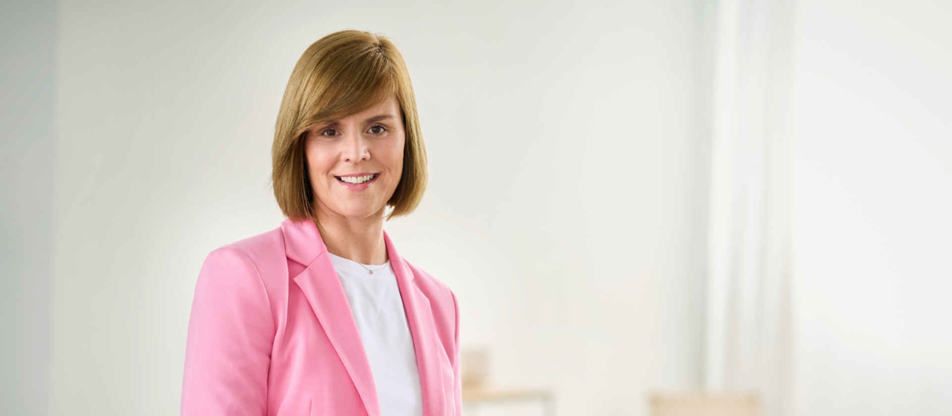 Eva Griewel, vrouw met roze blazer en wit hemd staat in een lichte ruimte.