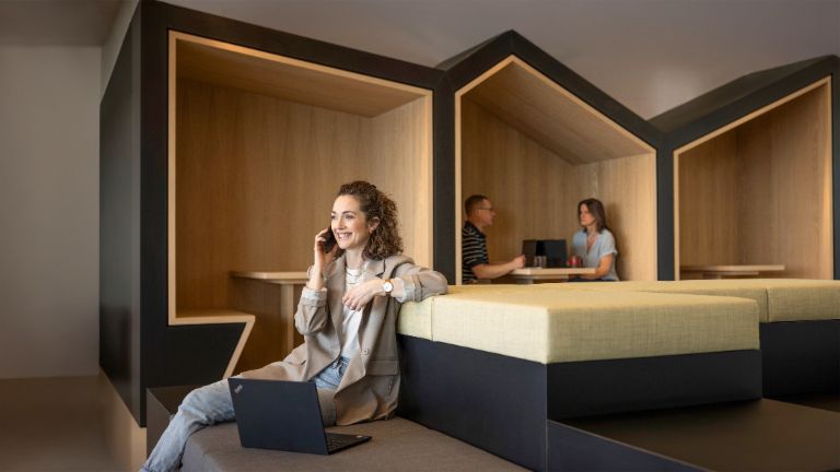 Een glimlachende persoon zit in een moderne kantoorlounge, telefoneert en werkt op een laptop. Op de achtergrond zijn twee andere mensen aan het werk.