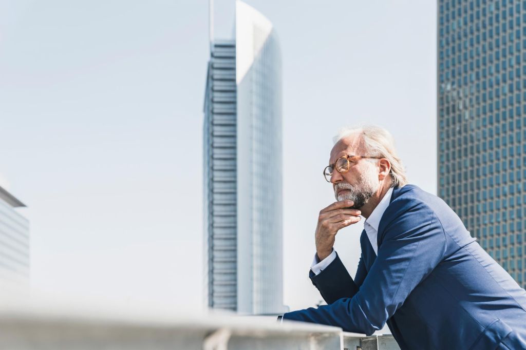 Man in suit leans against a parapet in front of office buildings and looks into the distance.