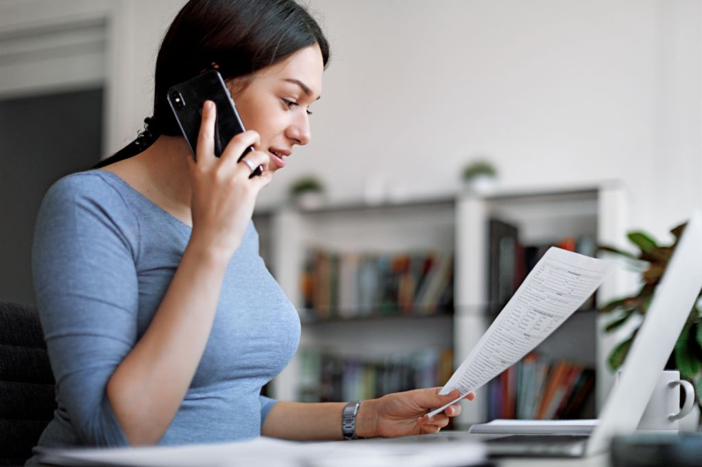 Vrouw in een blauwe top op de mobiele telefoon terwijl ze een factuur in haar handen houdt.