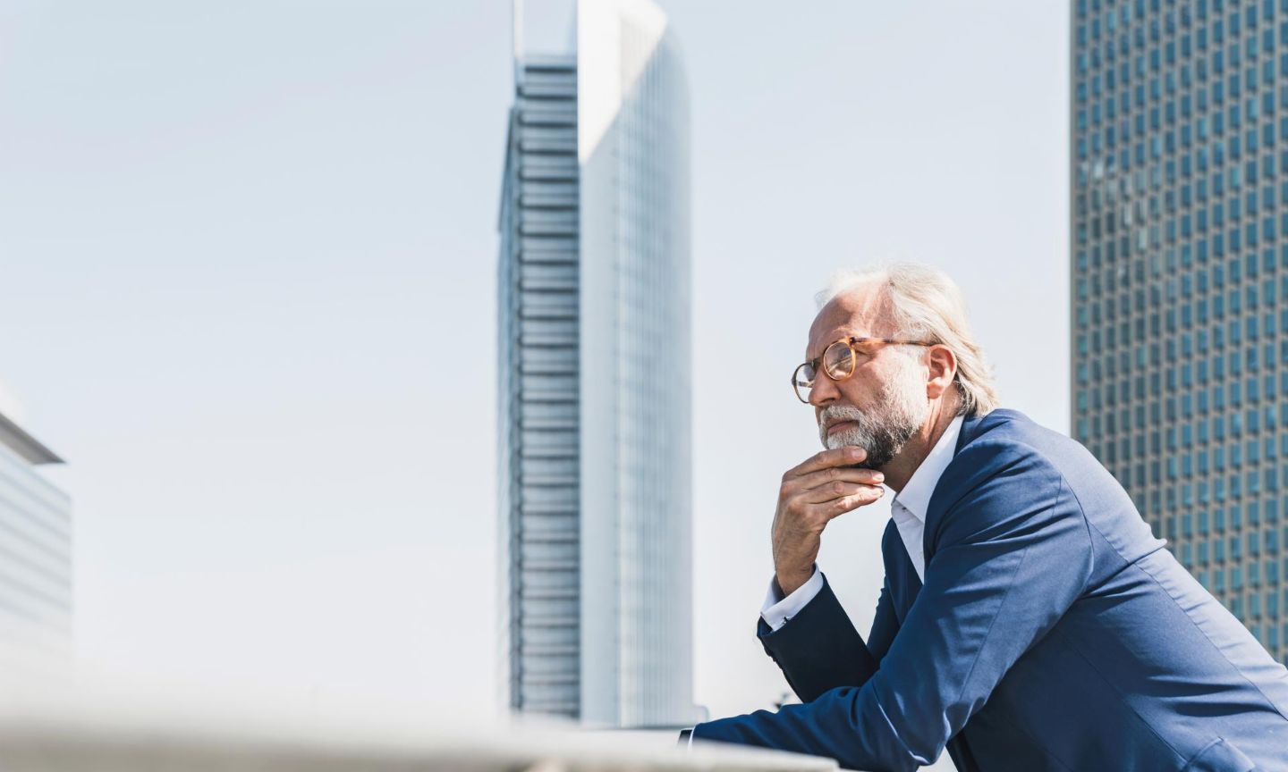 Man in suit leans against a parapet in front of office buildings and looks into the distance.